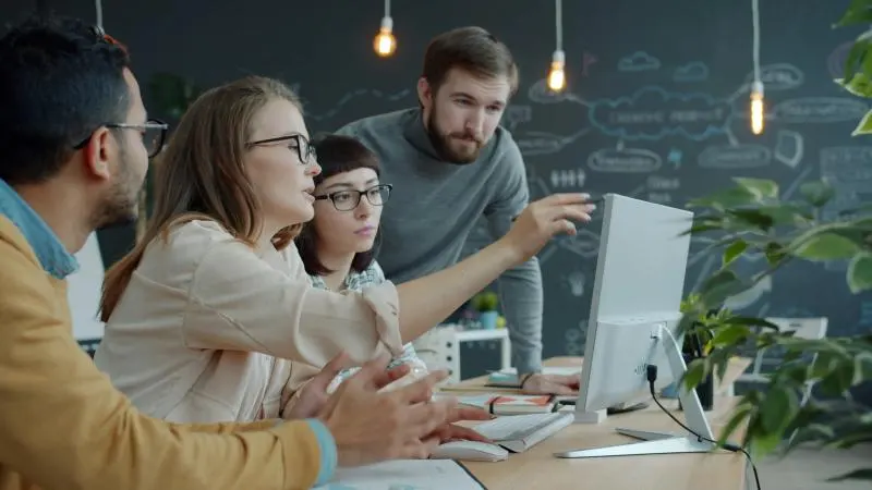 A small group of colleagues gathered around a desktop computer in a collaborative workspace, with one person pointing at the screen while others look on and discuss.