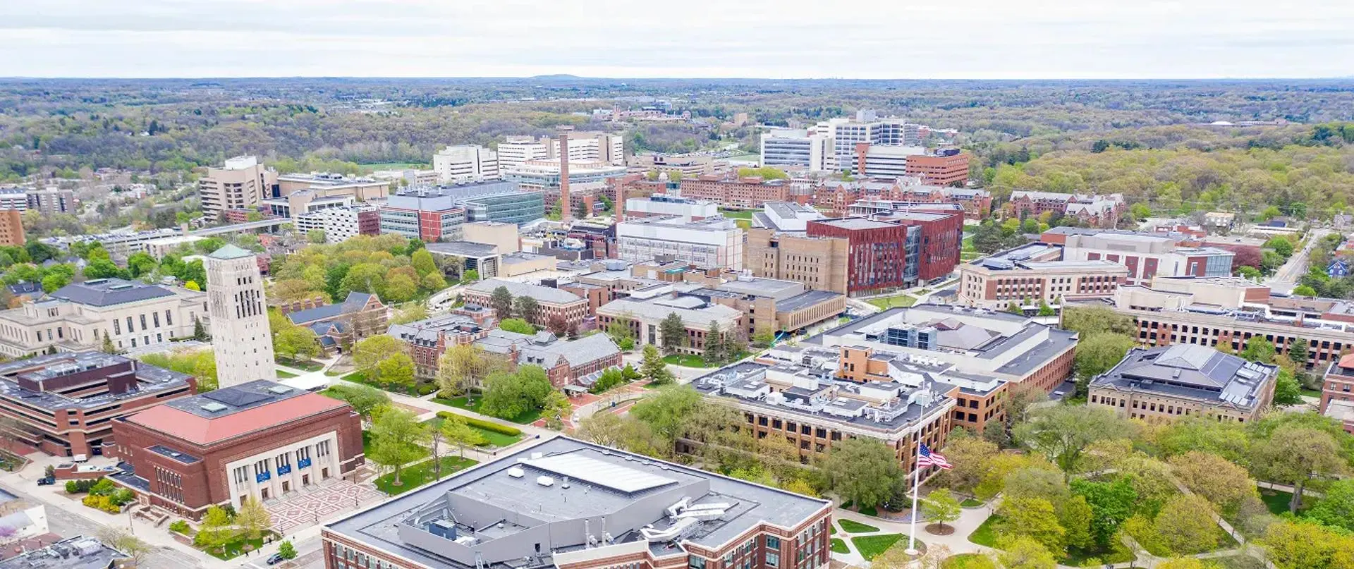 An aerial view of the University of Michigan's Ann Arbor campus in spring, showing a dense mix of historic red brick and modern academic buildings surrounded by trees, with the Burton Memorial Tower visible on the left.