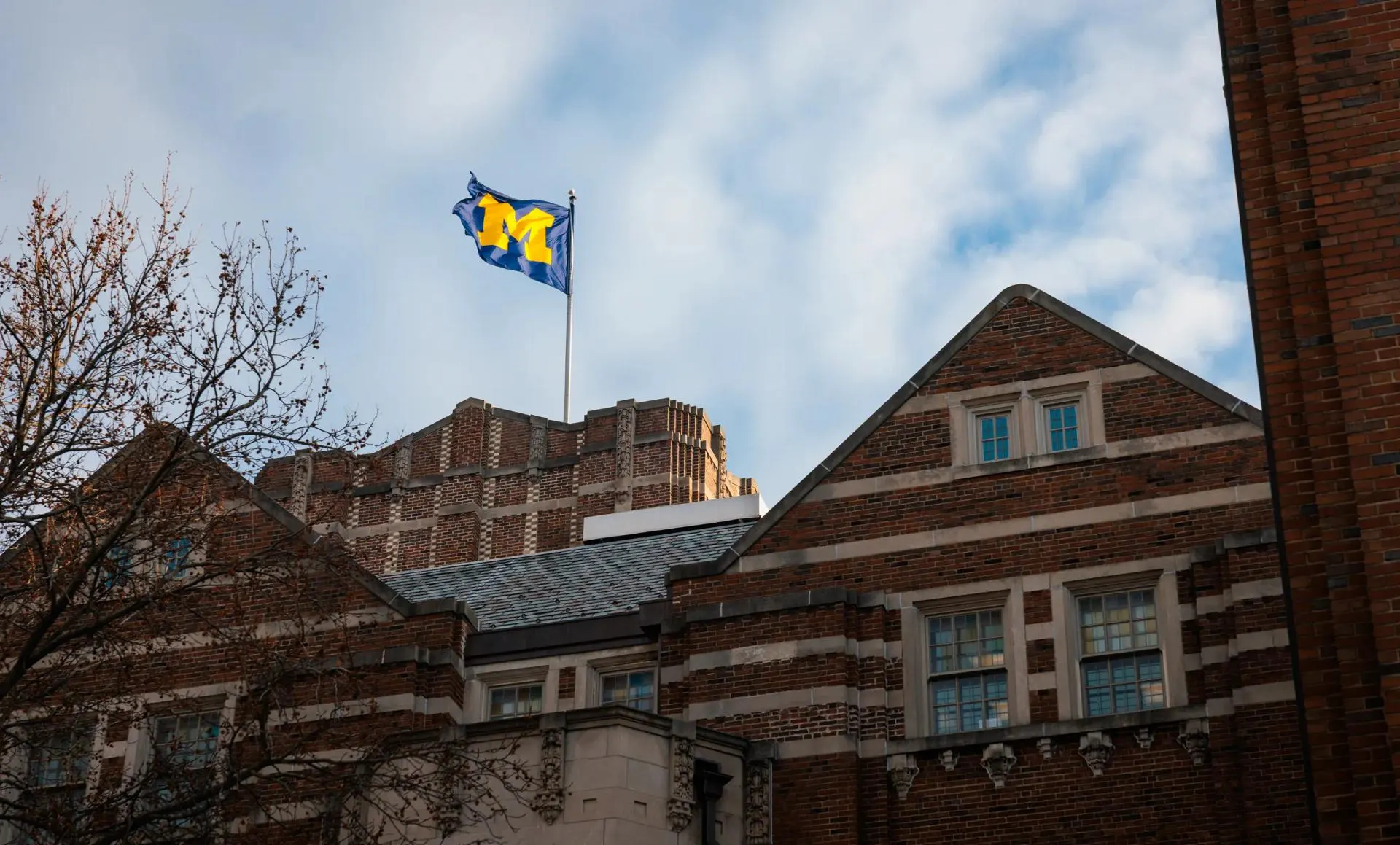 The University of Michigan flag, featuring a block M in maize on a navy background, flying above a red brick campus building against a partly cloudy sky.