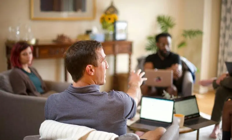 A small group of people seated in a living-room style setting, working on laptops and engaged in informal discussion, with one person speaking and gesturing toward the group.