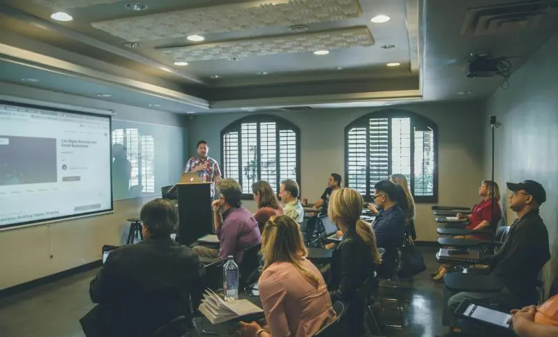 A presenter stands at a podium in front of a projected screen, addressing a room of approximately fifteen attendees seated at desks with notebooks and laptops.