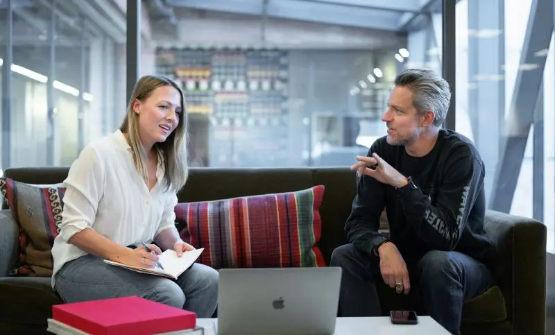 Two colleagues seated on a couch in a modern office lounge, engaged in conversation — one taking notes in a notebook while the other gestures while speaking, with a laptop open on the table between them.