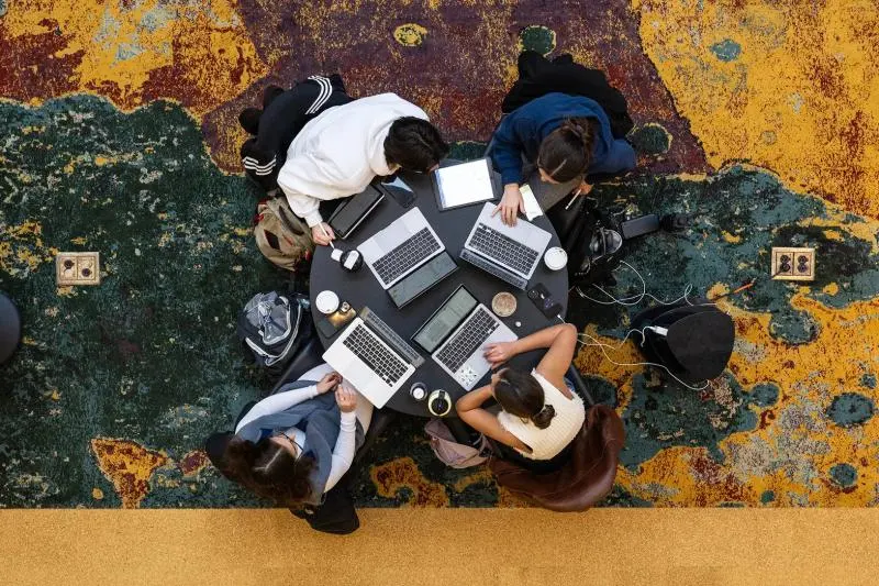 An overhead view of four people seated around a small round table, each working on a laptop, with bags and accessories spread around them on a colorful, textured floor.